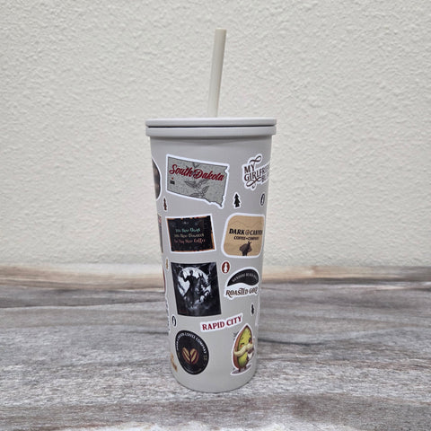 A gray insulated tumbler with a white lid and straw, decorated with multiple travel-themed stickers including 'South Dakota', 'Rapid City', set on a gray granite surface against a textured off-white wall.
